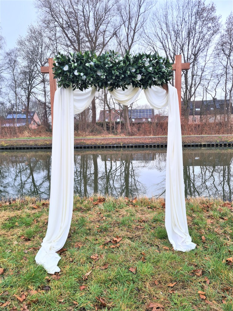 Vooraanzicht White Dreams ceremonieboog. Witte bloemen met groen er rond en witte doeken op een houten boog. 